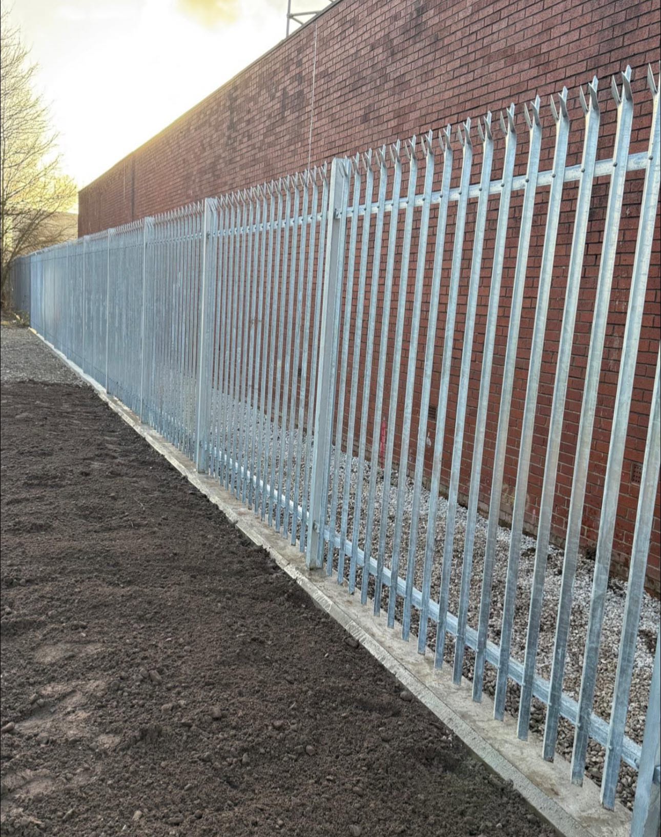 Galvanised palisade fencing installed alongside a red brick industrial wall on a concrete plinth