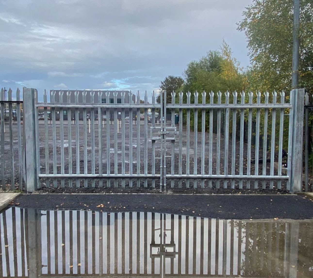 Galvanised palisade double gates with drop-bolt centre stop reflected in a puddle on a commercial yard